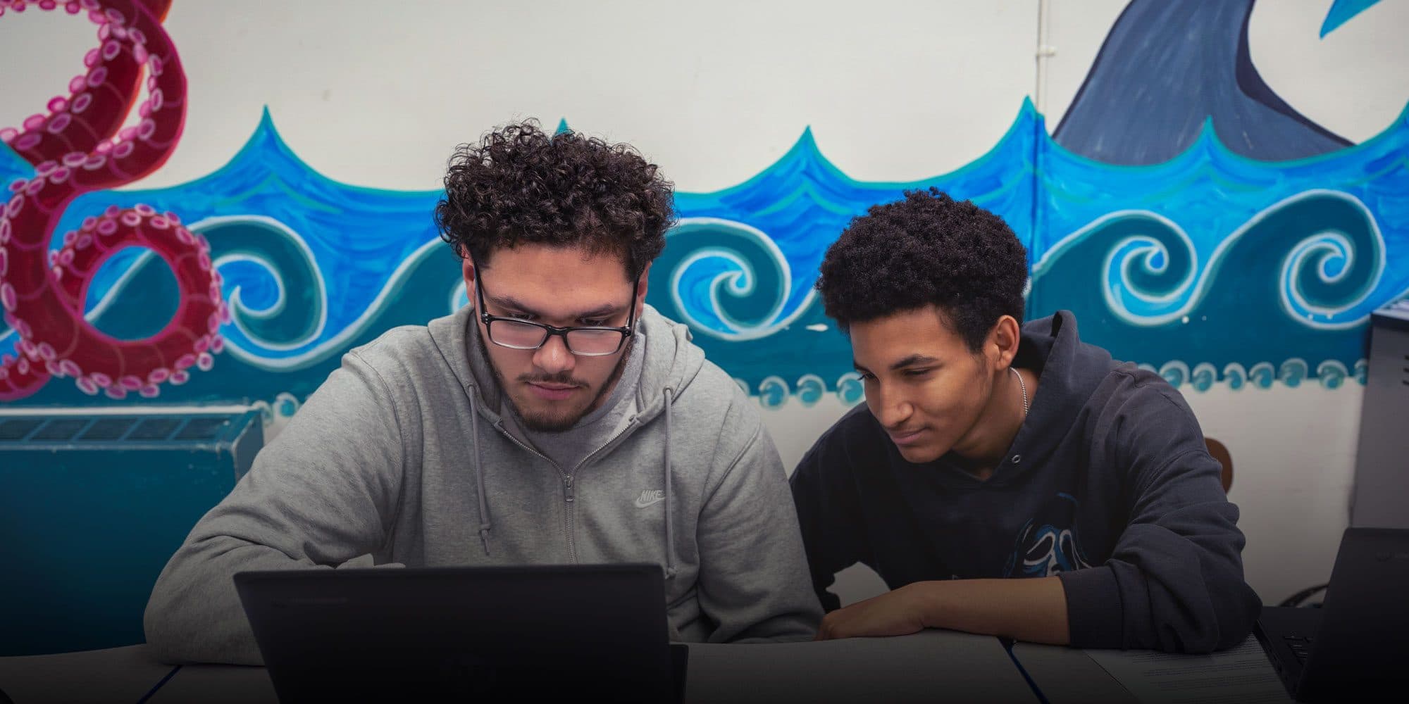 Two students working together on a laptop in front of an ocean mural.